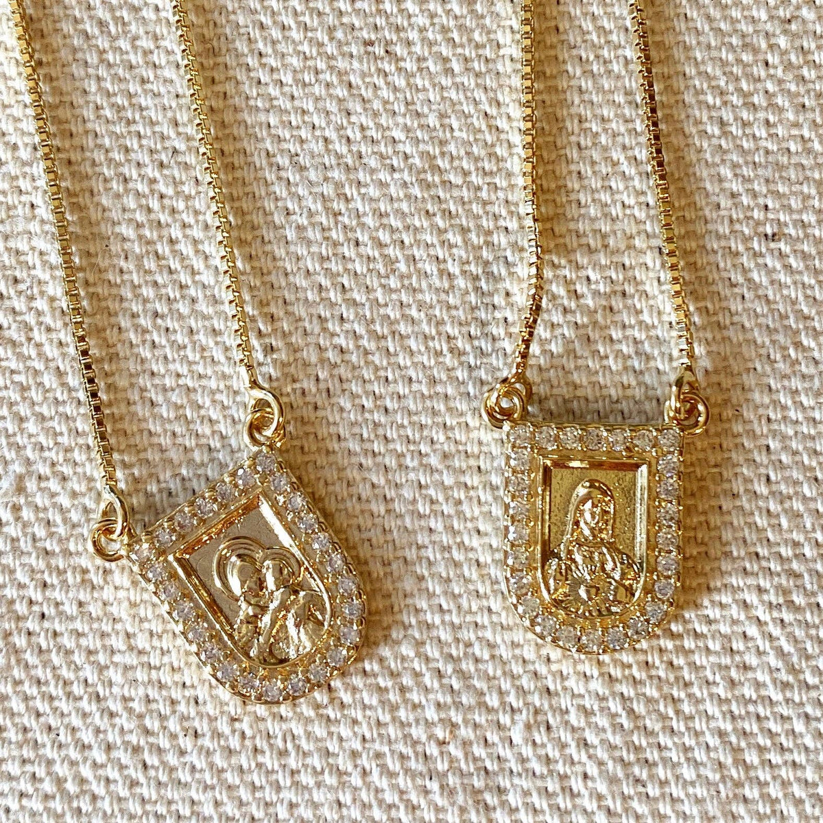 Two gold necklaces with religious-themed pendants on a textured beige background