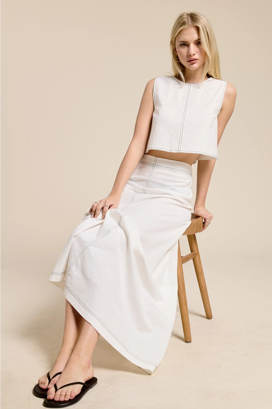 Woman wearing a white sleeveless top and skirt sitting on a wooden stool against a beige background