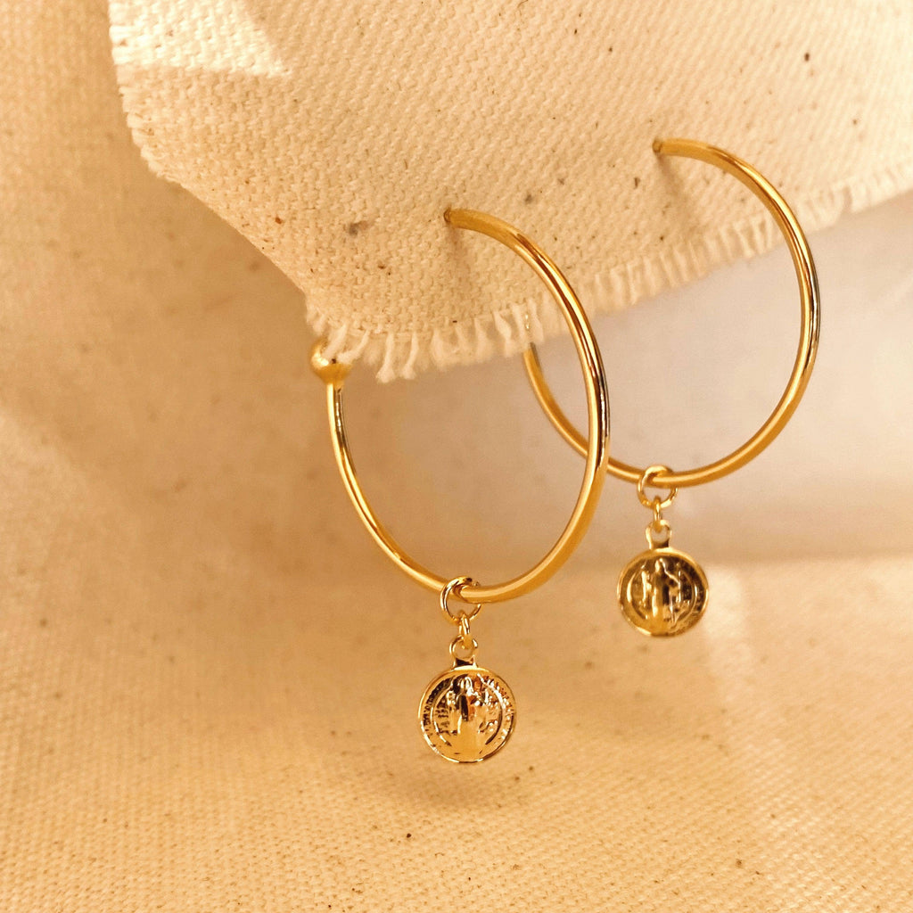 A pair of gold-tone hoop earrings with small dangling textured round charms, displayed on a beige fabric surface.