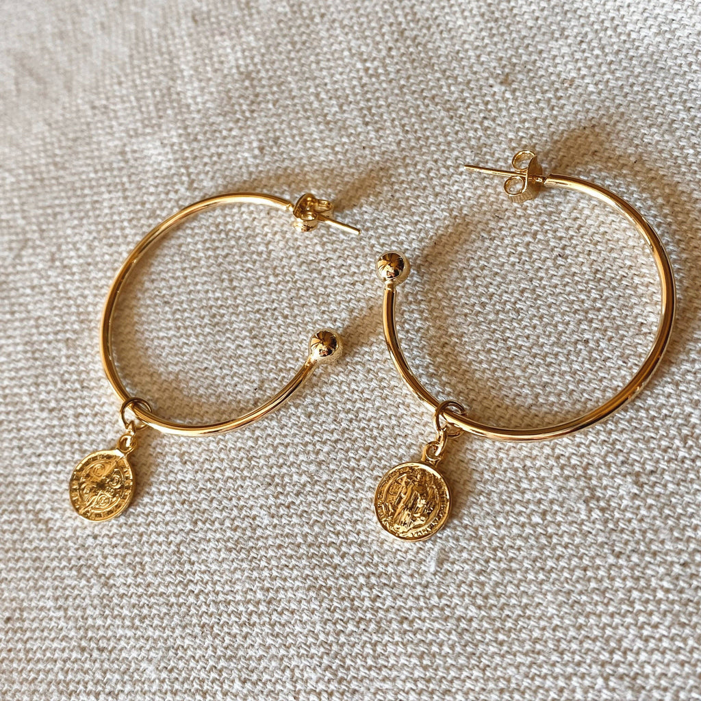 A pair of gold hoop earrings with small coin-style charms, displayed on a textured beige fabric background.