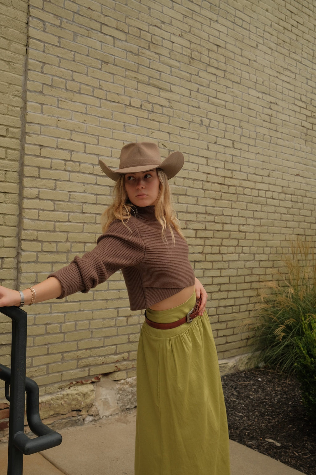 A woman wearing a brown ribbed long-sleeve crop top, olive green maxi skirt, and brown cowboy hat, leaning against a black metal frame in front of a beige brick wall.