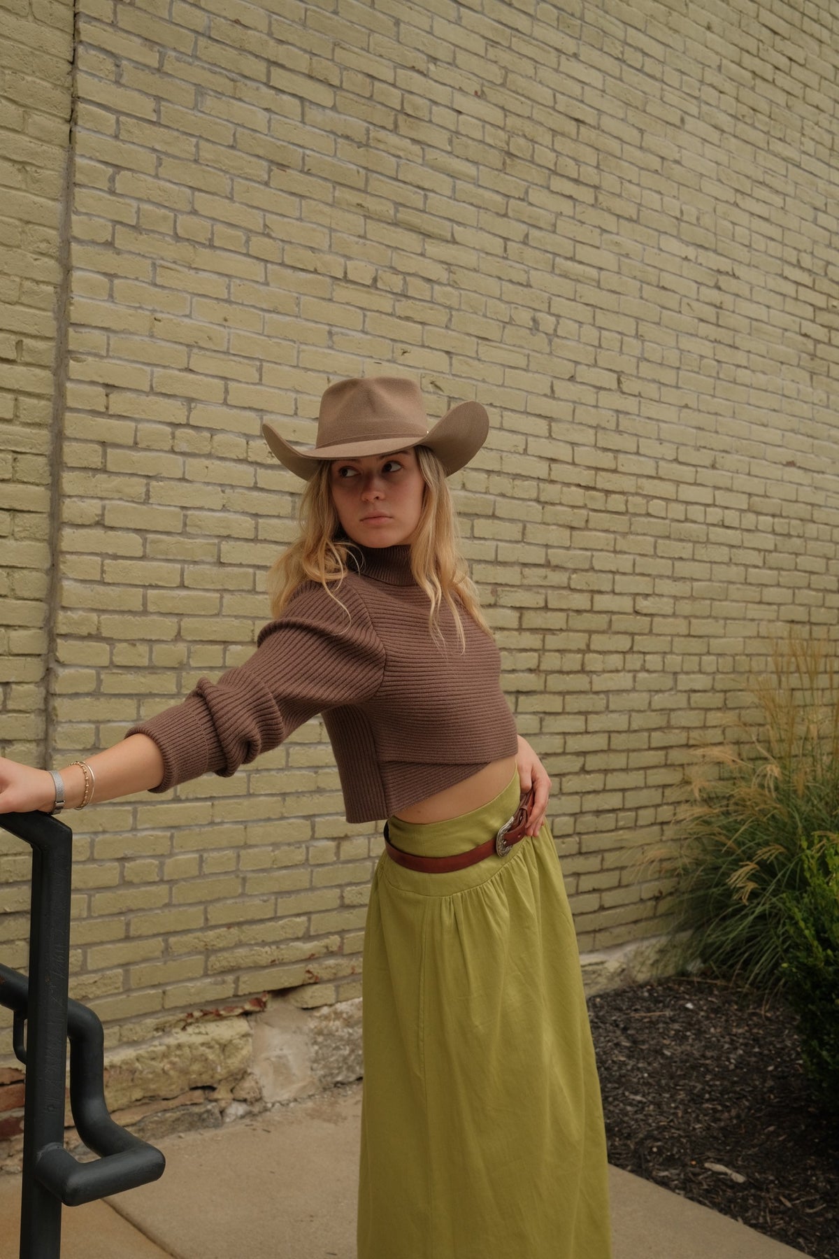 A woman wearing a brown ribbed long-sleeve crop top, olive green maxi skirt, and brown cowboy hat, leaning against a black metal frame in front of a beige brick wall.