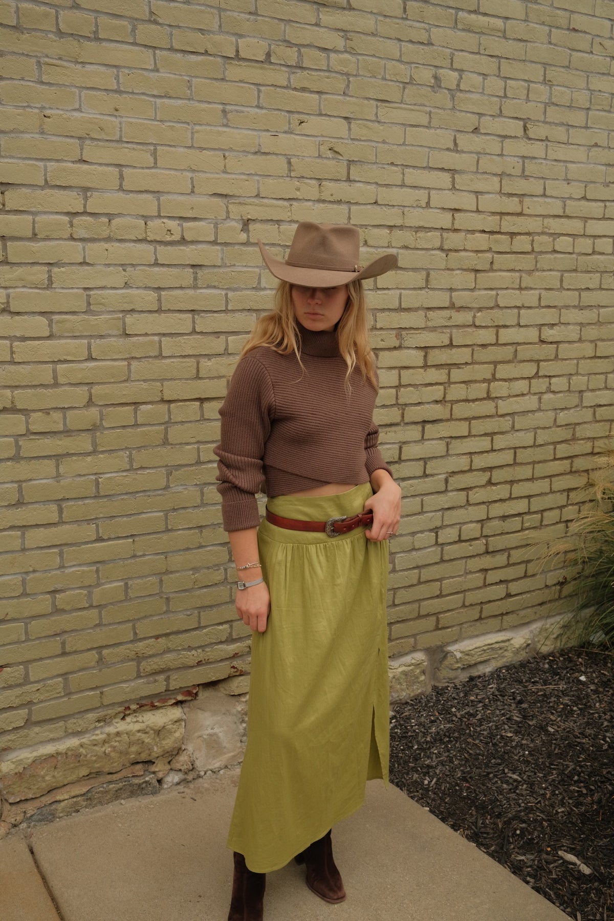 A woman wearing a brown knit turtleneck, olive green skirt, brown belt, and brown cowboy hat, standing against a beige brick wall.