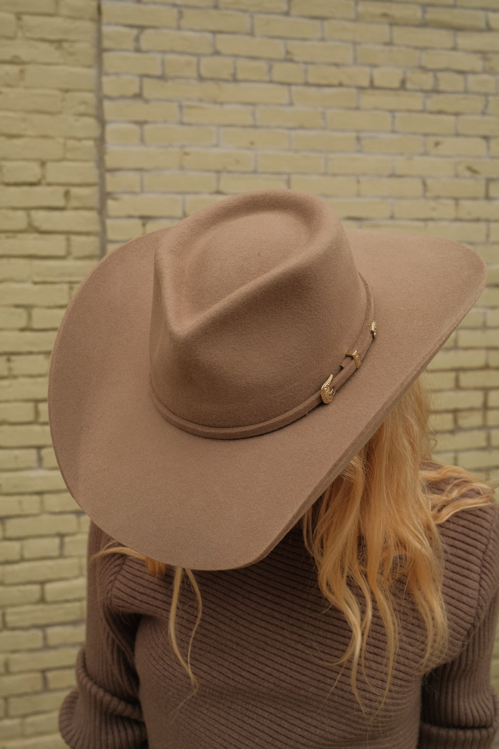 A person wearing a wide-brimmed tan felt cowboy hat with a small gold-toned decorative band, standing in front of a light brick wall.
