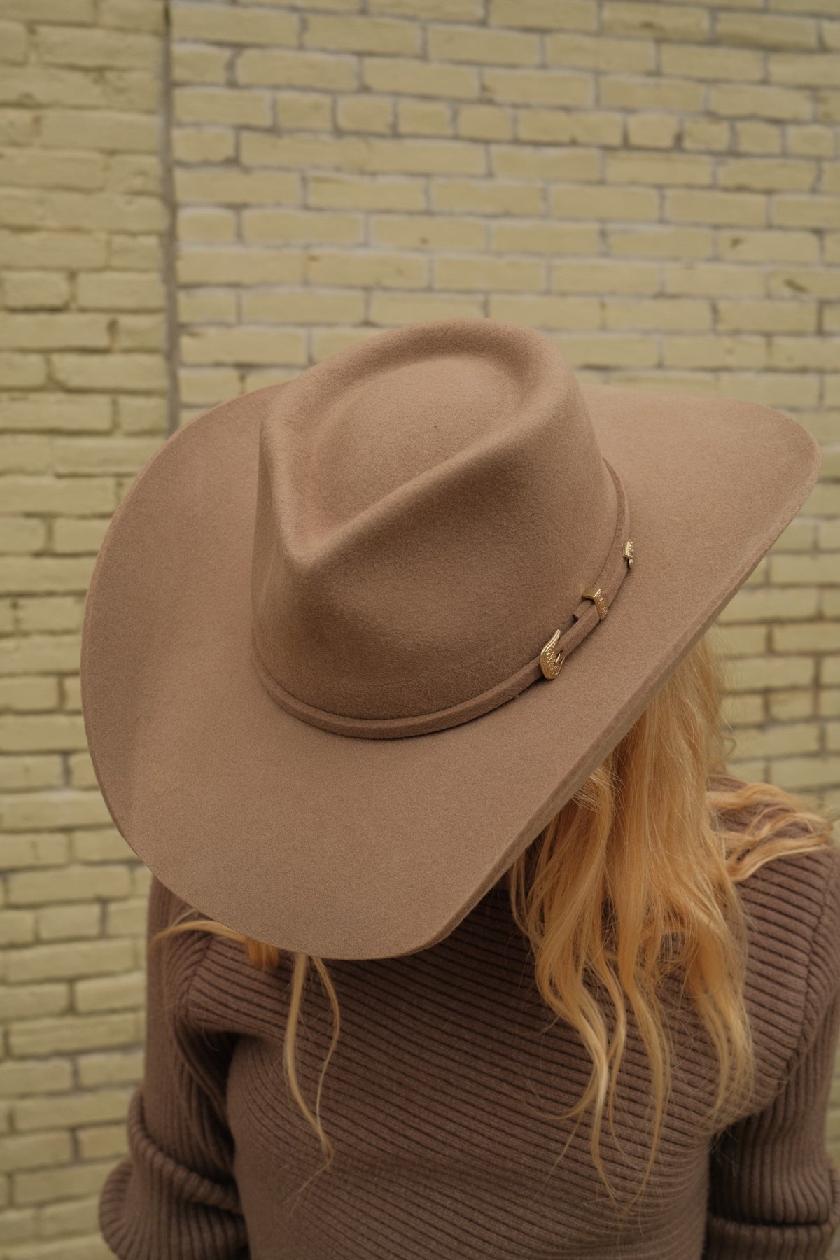 A person wearing a wide-brimmed tan felt cowboy hat with a small gold-toned decorative band, standing in front of a light brick wall.