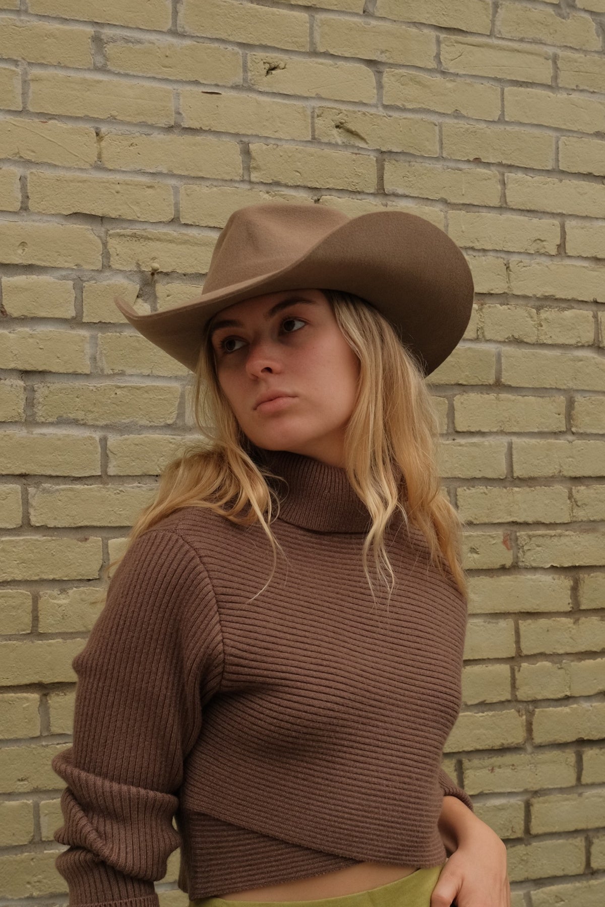 A woman wearing a brown ribbed turtleneck sweater and a brown cowboy hat, standing against a light beige brick wall.