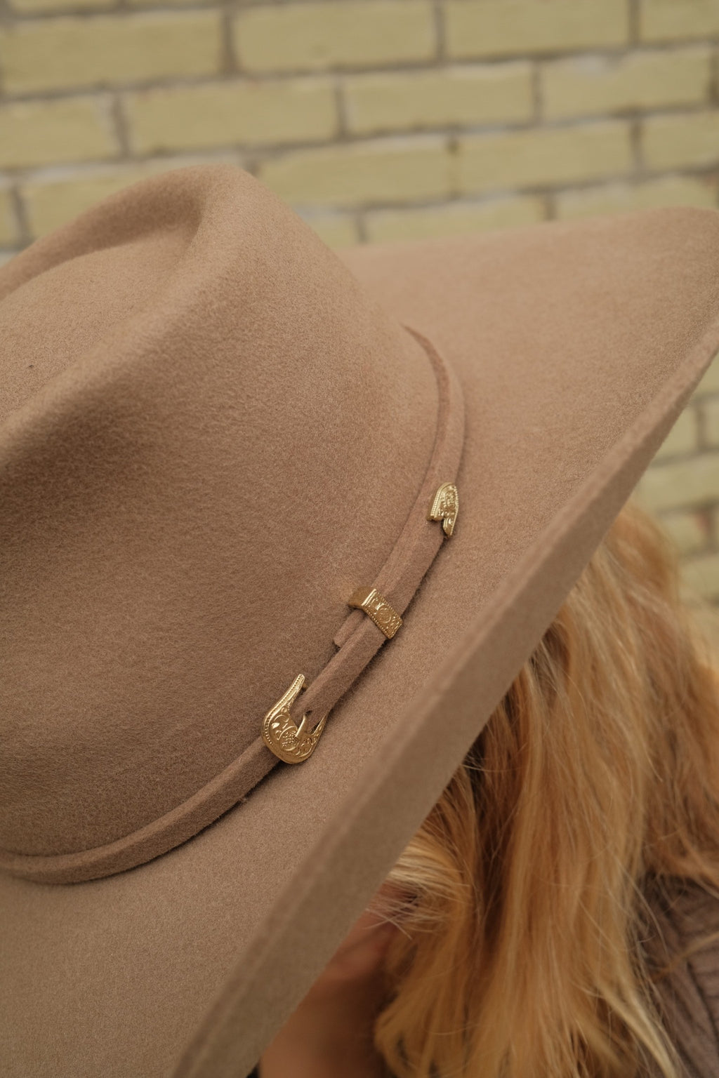 A close-up of a wide-brimmed felt cowboy hat in a warm tan-brown color, featuring a matching band with gold-toned decorative hardware, worn by a person with blonde hair against a light brick wall.