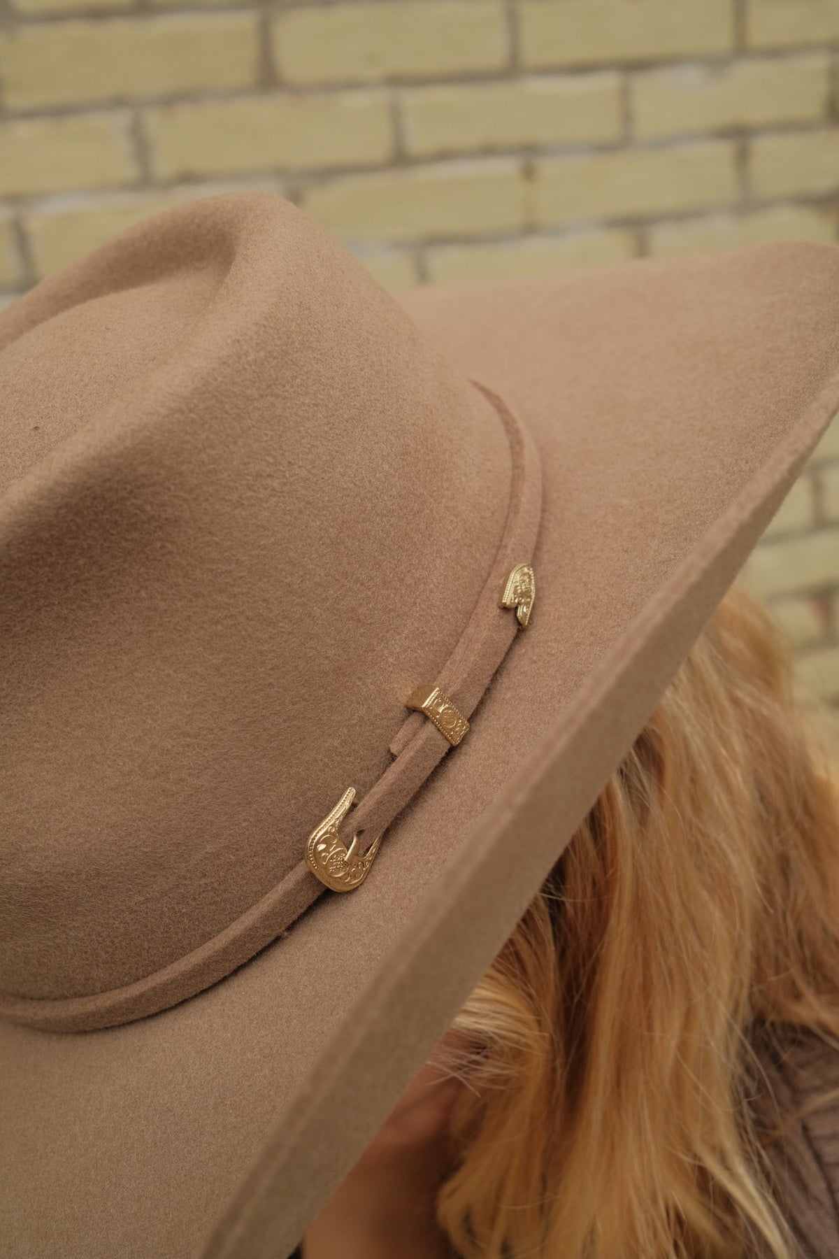 A close-up of a wide-brimmed felt cowboy hat in a warm tan-brown color, featuring a matching band with gold-toned decorative hardware, worn by a person with blonde hair against a light brick wall.