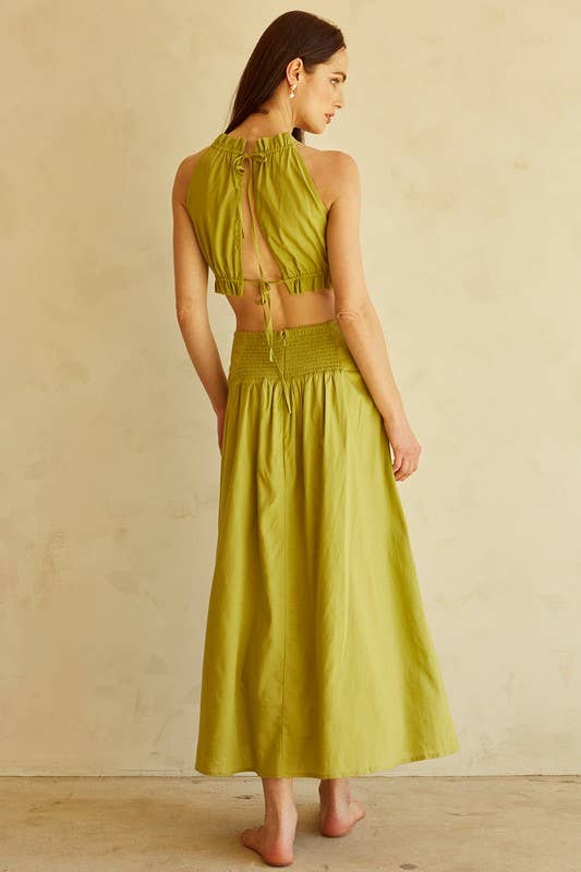 A woman stands barefoot against a textured beige wall, wearing a bright yellow-green two-piece set with a halter crop top and a matching high-waisted maxi skirt.