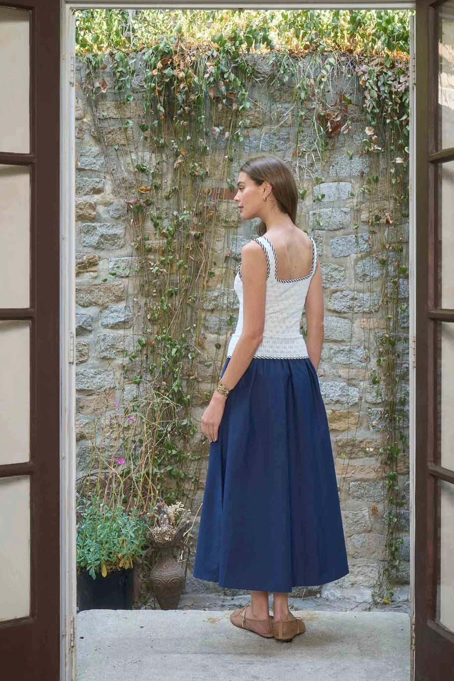 Woman in a white top and blue skirt standing in a doorway with a stone wall and plants in the background