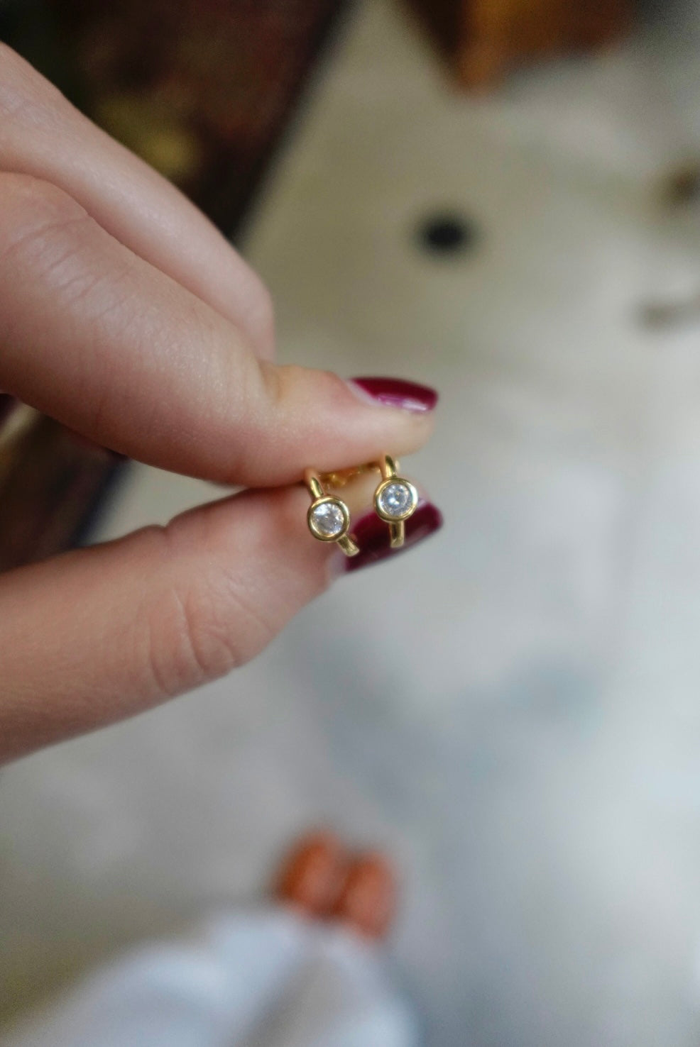 Gold earrings with clear stones held between fingers against a blurred background
