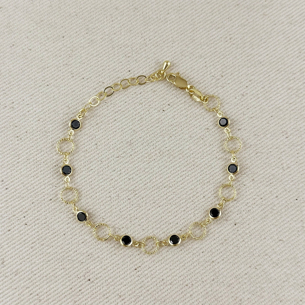 A delicate gold chain bracelet with alternating textured open circles and small round black gemstone accents, displayed on a textured beige fabric background.