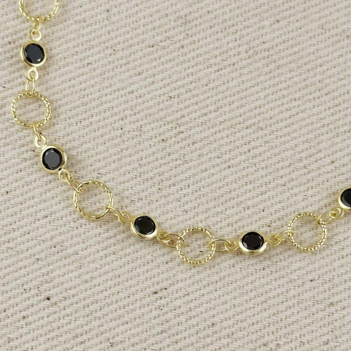 A gold chain bracelet featuring alternating smooth oval and textured circular links, each set with a small round black onyx stone, displayed on a textured beige fabric background.