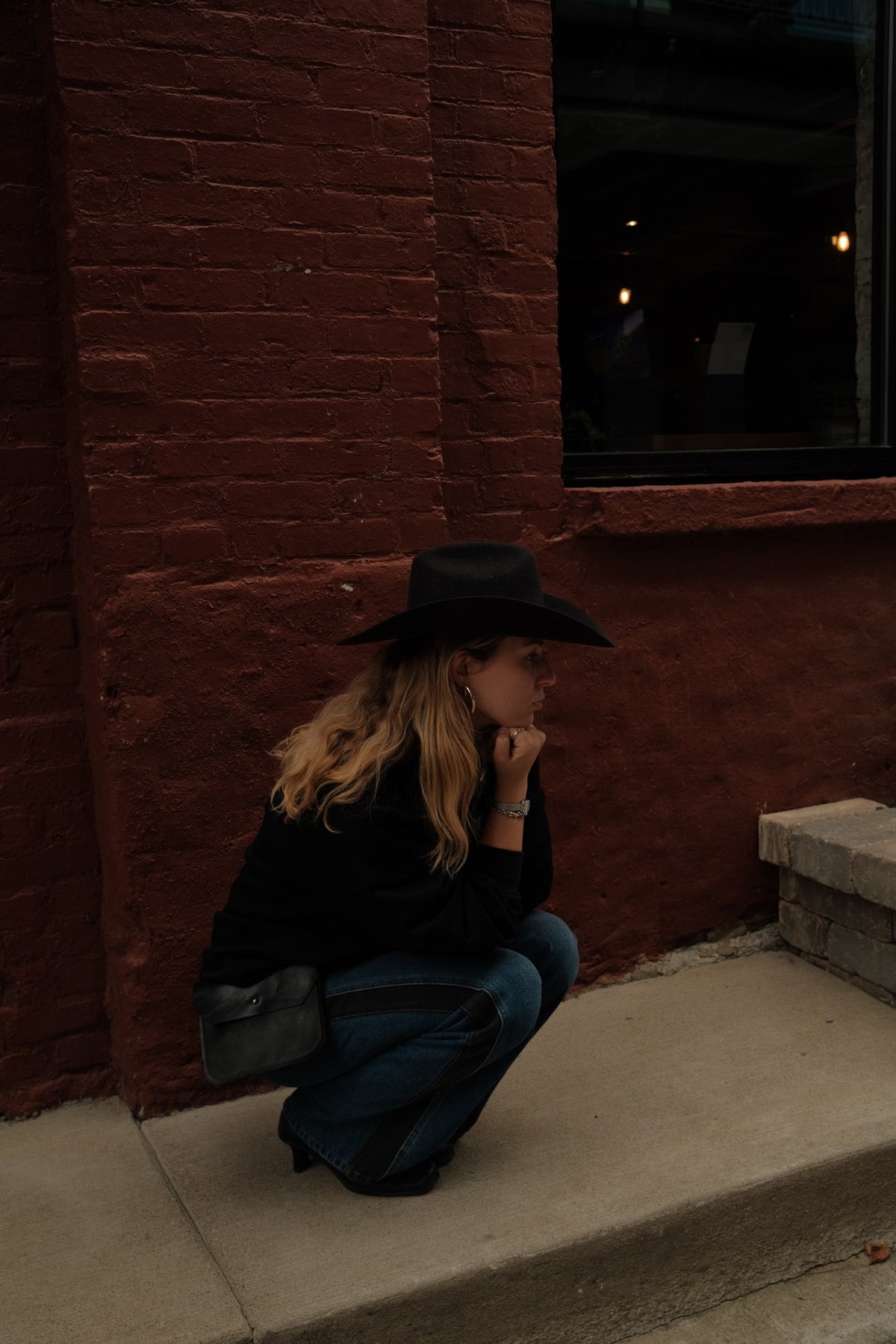 A woman wearing a black wide-brim hat, black top, and blue jeans crouches on concrete steps against a red brick building wall.