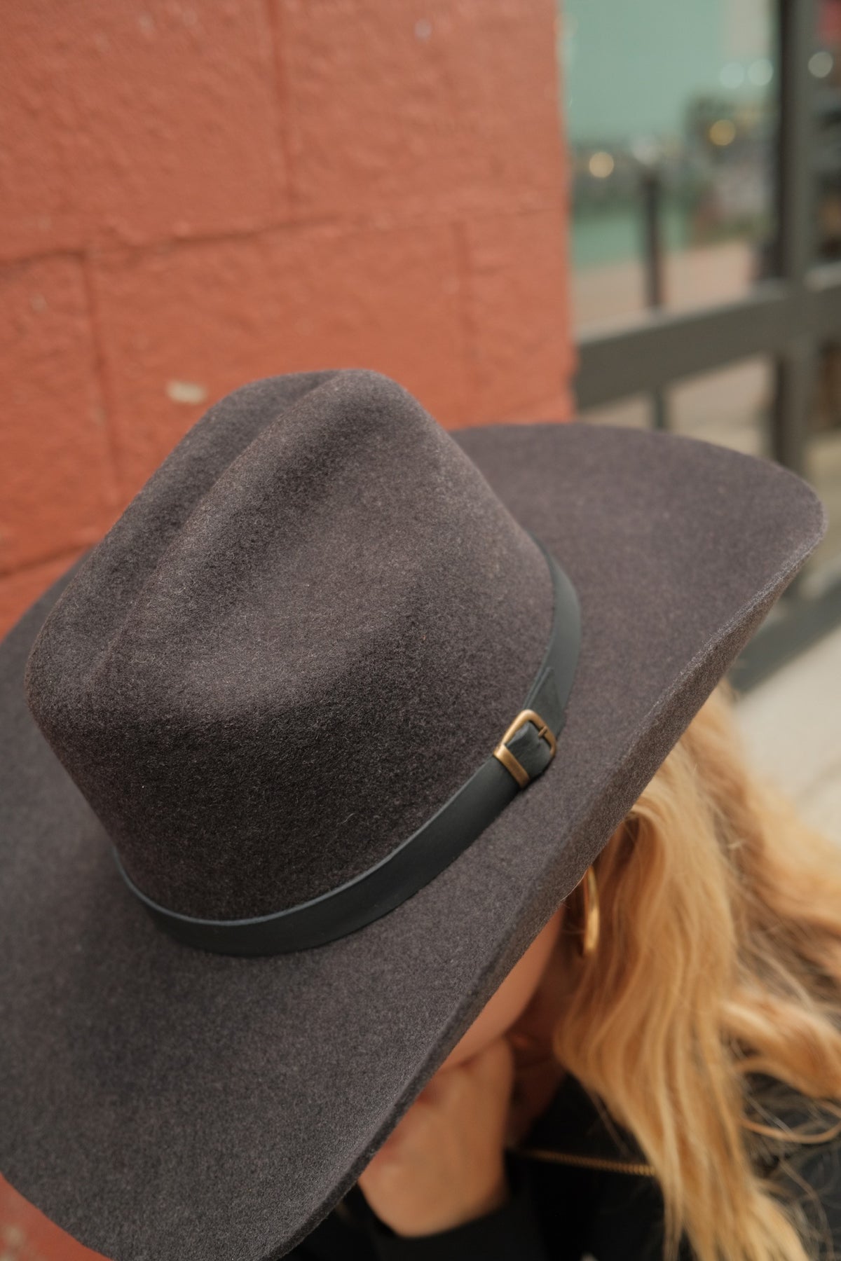 A close-up shot of a dark gray felt cowboy hat with a black band and gold buckle, worn by a person with blonde hair visible in the background against a brick wall.