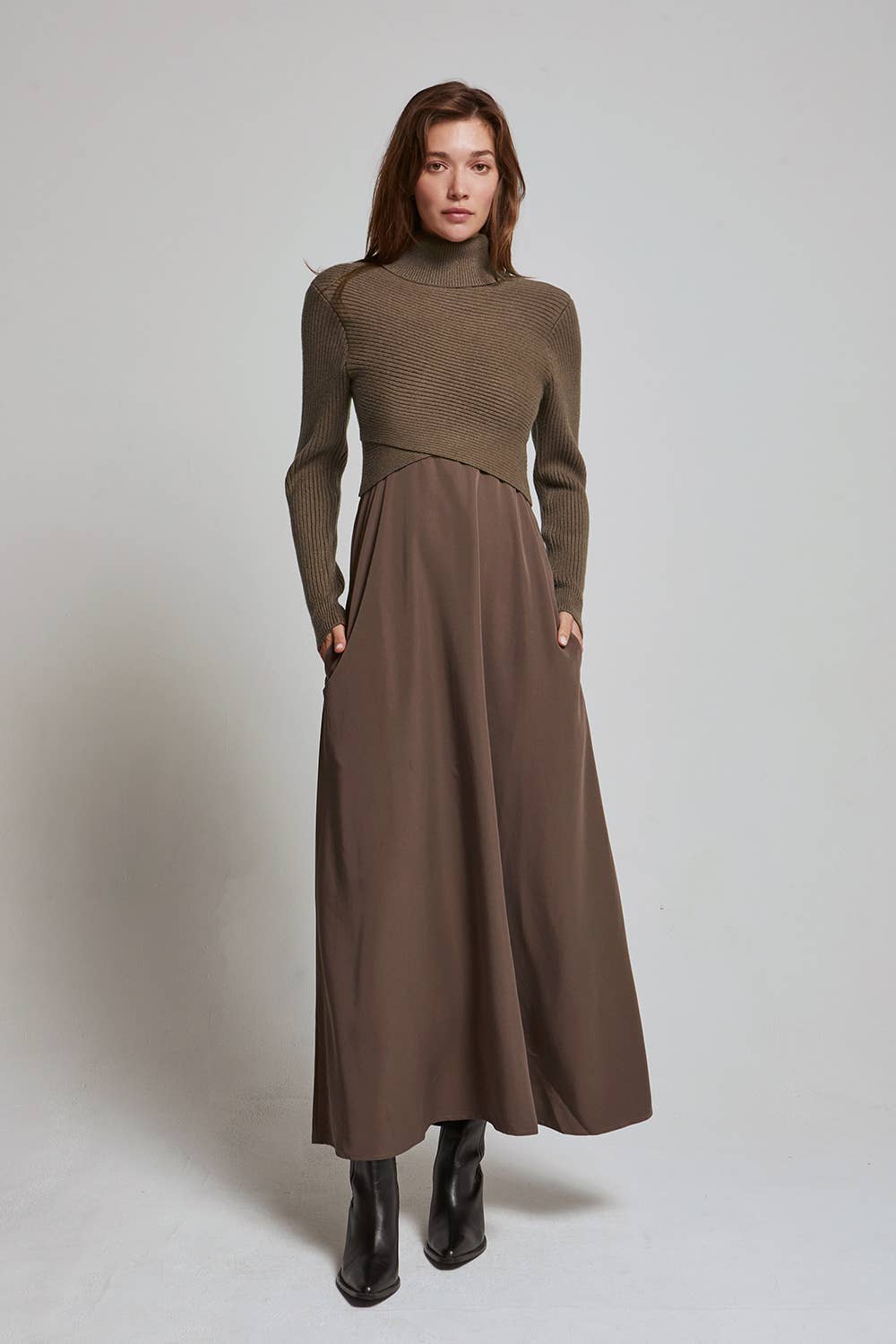 A woman modeling a long-sleeve, high-neck, two-tone dress in earthy brown tones, featuring a fitted ribbed top and a flowing skirt with side pockets, paired with black ankle boots, against a neutral studio backdrop.