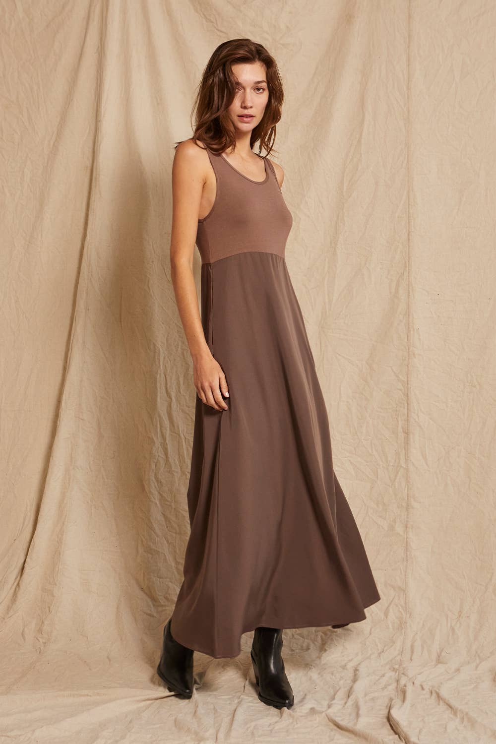 A woman modeling a sleeveless, scoop-neck maxi dress in a solid taupe-brown color, paired with black ankle boots, standing against a draped beige fabric backdrop.