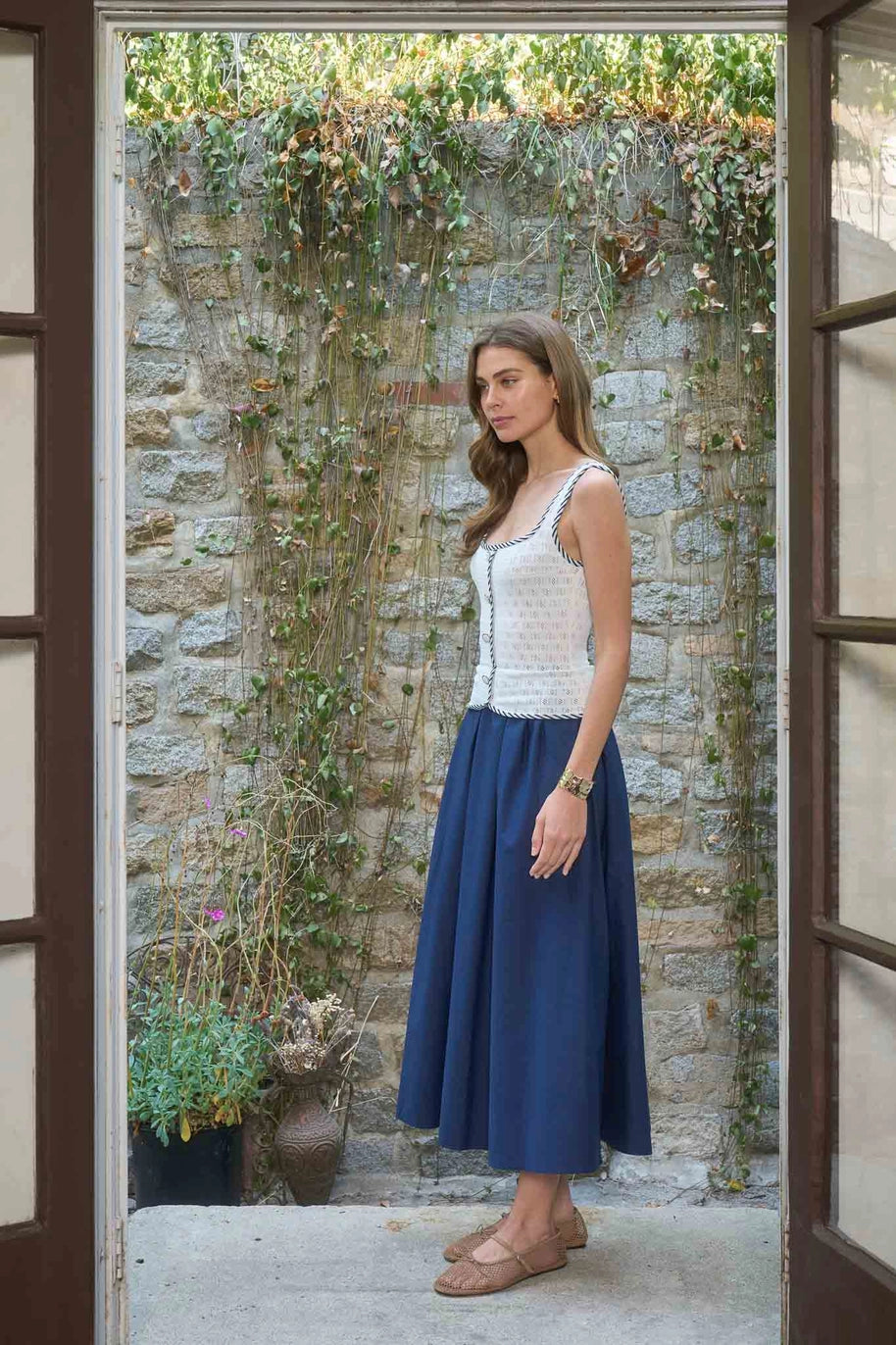 Woman in a white top and blue skirt standing in front of a stone wall with greenery.