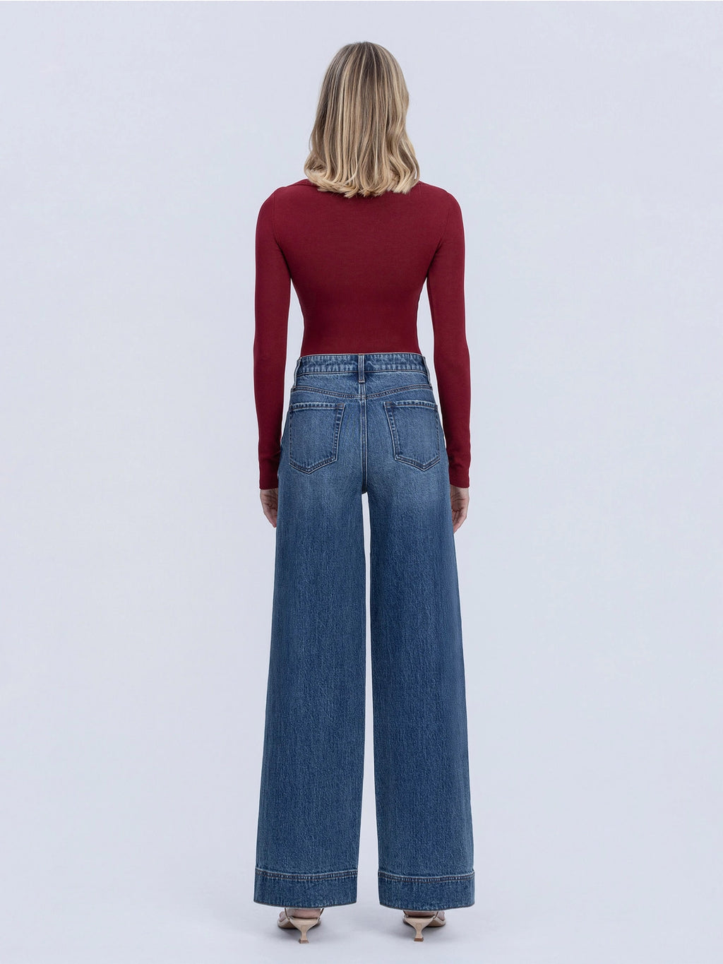 A woman viewed from the back wearing a fitted burgundy long-sleeve top and high-waisted wide-leg blue jeans against a light gray studio background.