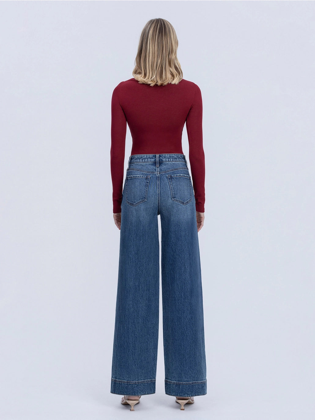 A woman viewed from the back wearing a fitted burgundy long-sleeve top and high-waisted wide-leg blue jeans against a light gray studio background.