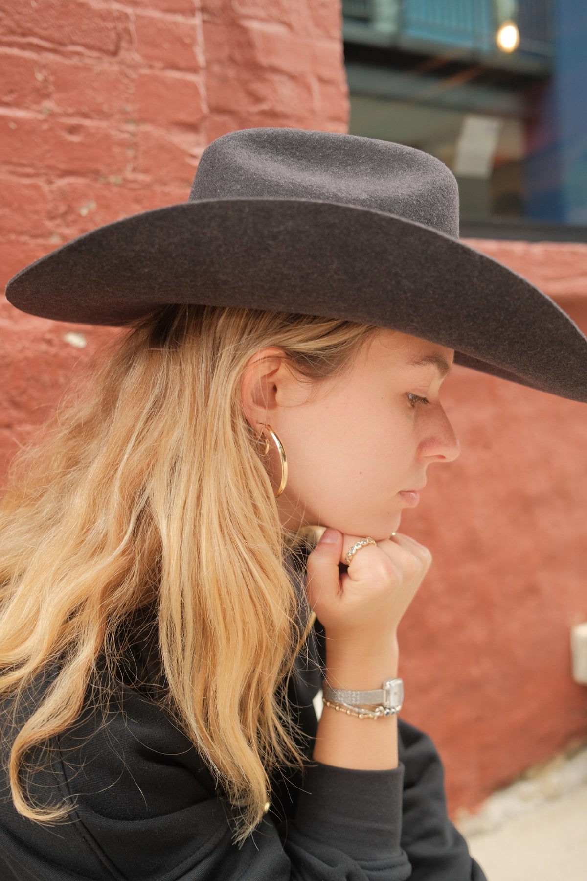 A woman in profile wearing a wide-brimmed dark gray felt cowboy hat, gold hoop earrings, and a silver watch, standing against a red brick wall.