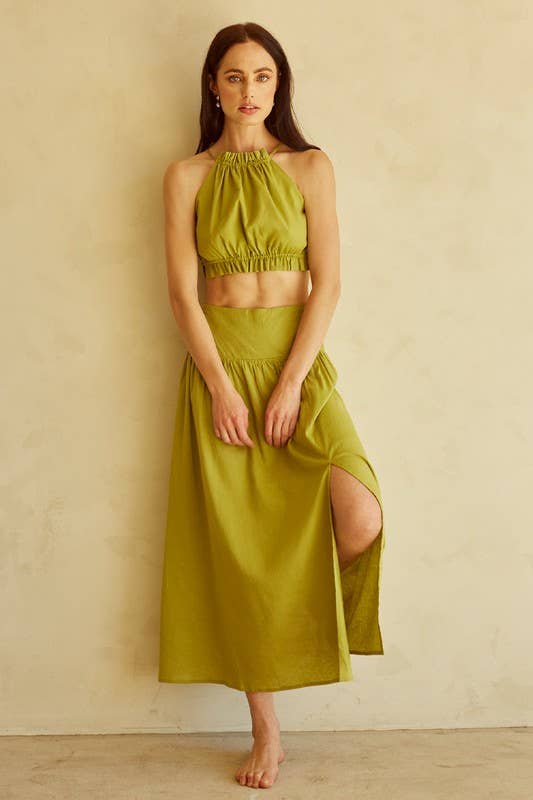 A woman stands barefoot against a warm beige textured wall wearing a matching two-piece olive green set consisting of a halter crop top and a high-slit midi skirt.