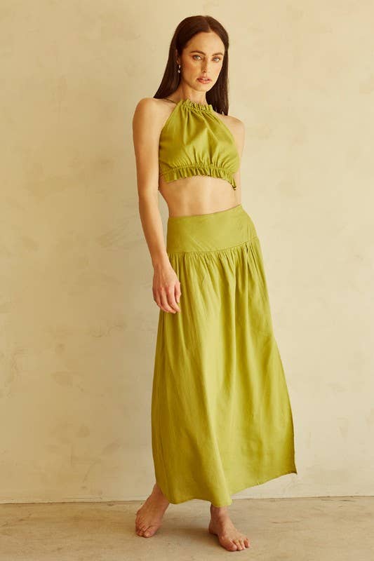 A woman modeling a matching olive green two-piece set consisting of a halter crop top and a high-waisted maxi skirt, standing against a textured beige wall.