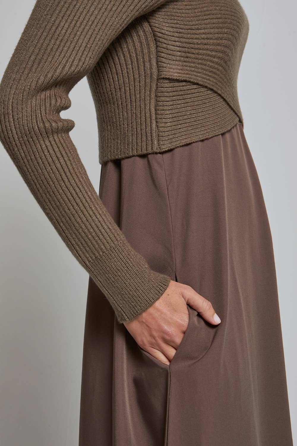 Close-up of a person wearing a ribbed cropped knit sweater in taupe-brown and a matching skirt, with one hand in a pocket, against a plain light gray background.