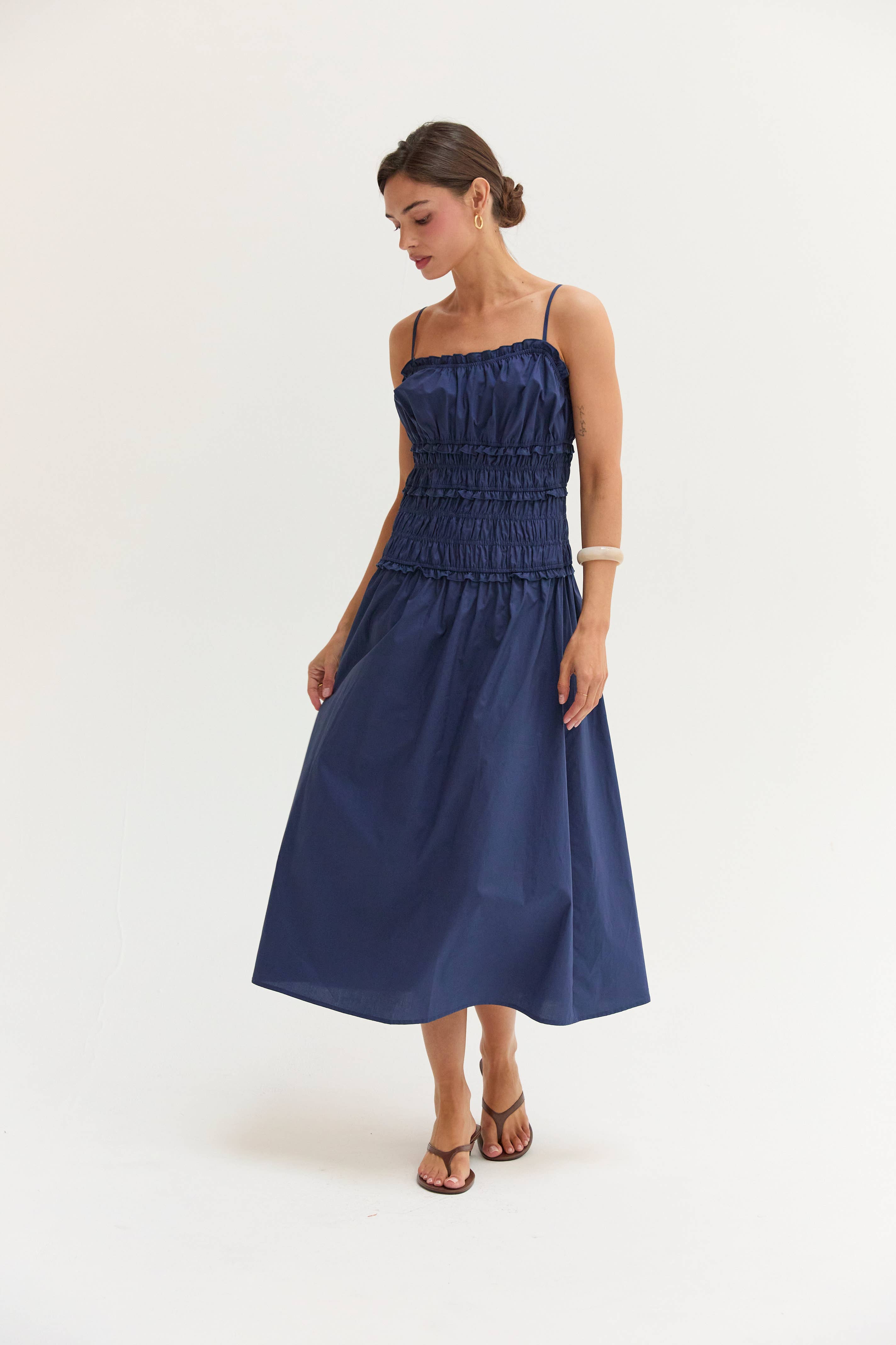 A woman modeling a navy blue midi dress with thin straps, a smocked bodice, and a flared skirt, standing against a plain white studio background.