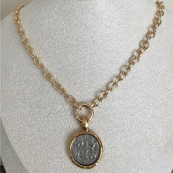 A gold chain necklace with a round pendant featuring an embossed cross, displayed on a white fabric jewelry bust.