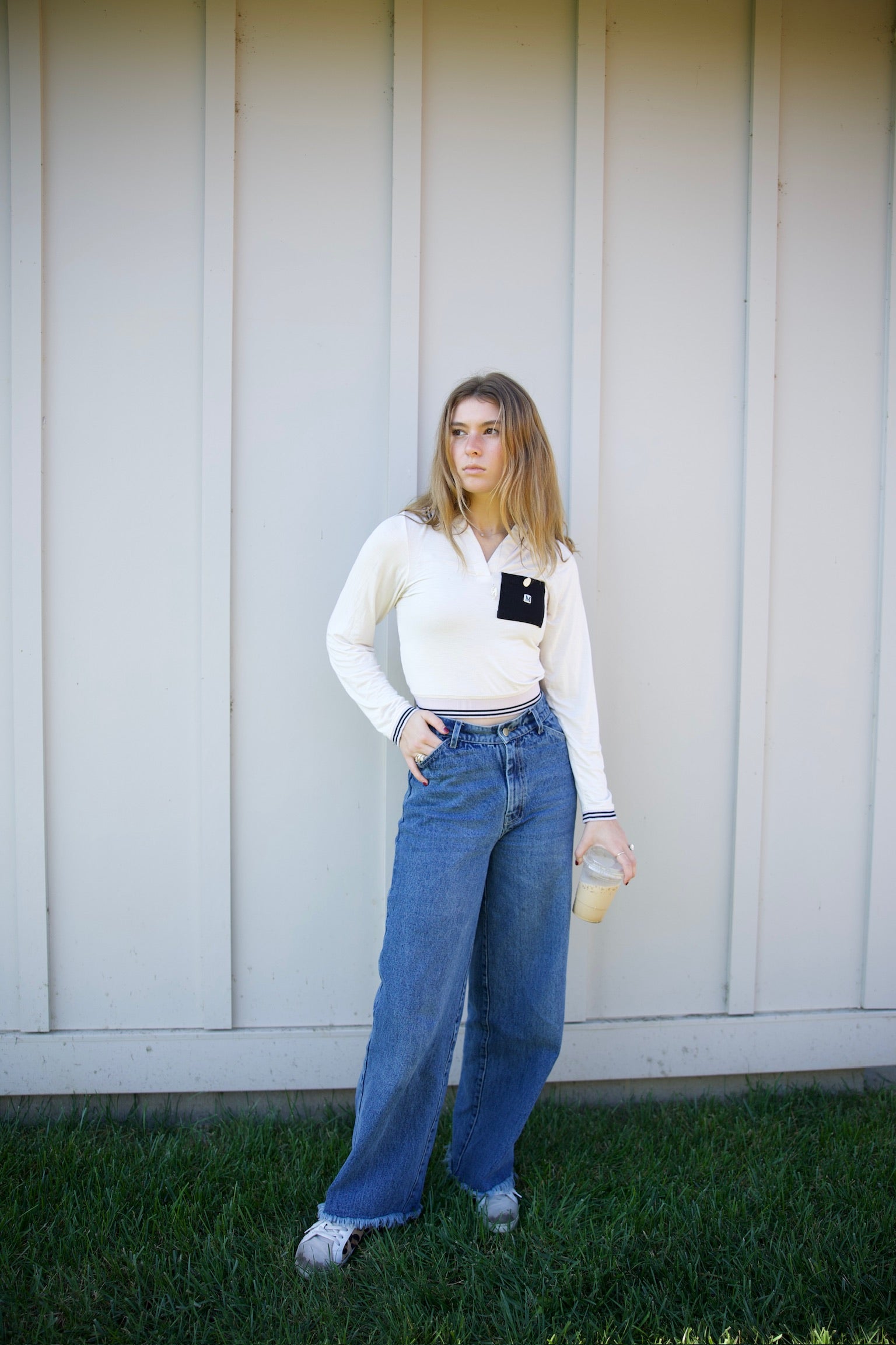 Person wearing a white blouse with a black patch and blue jeans standing against a light-colored wall.