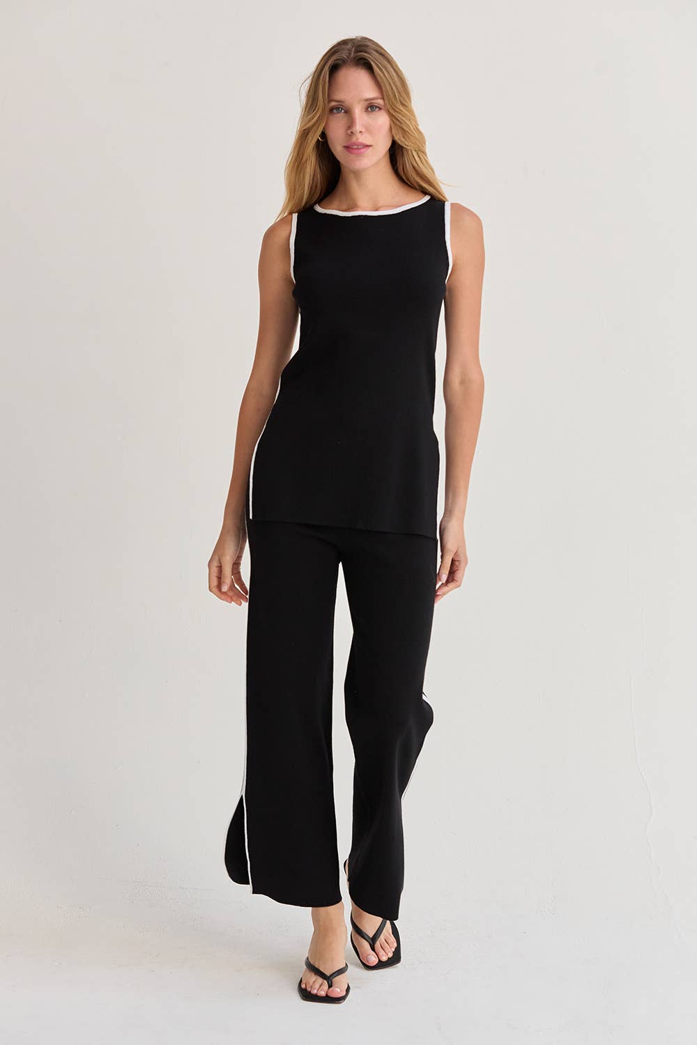 A woman modeling a black sleeveless knit top and matching wide-leg pants set, featuring a white contrast trim at the neckline and along the sides of the top, against a plain light gray studio background.