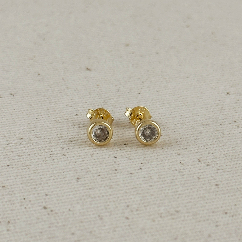 Gold stud earrings with clear gemstones on a light gray fabric background