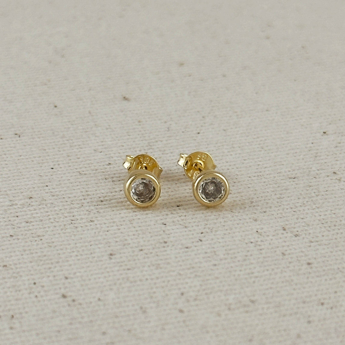 Gold stud earrings with clear gemstones on a light gray fabric background