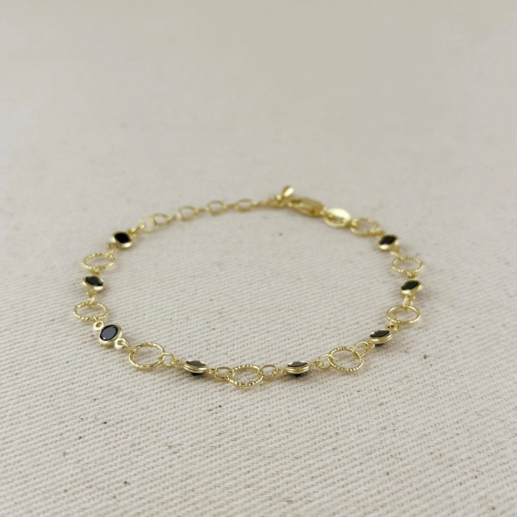 A delicate gold chain bracelet with small oval links and a few dark oval stone accents, displayed on a textured beige fabric background.