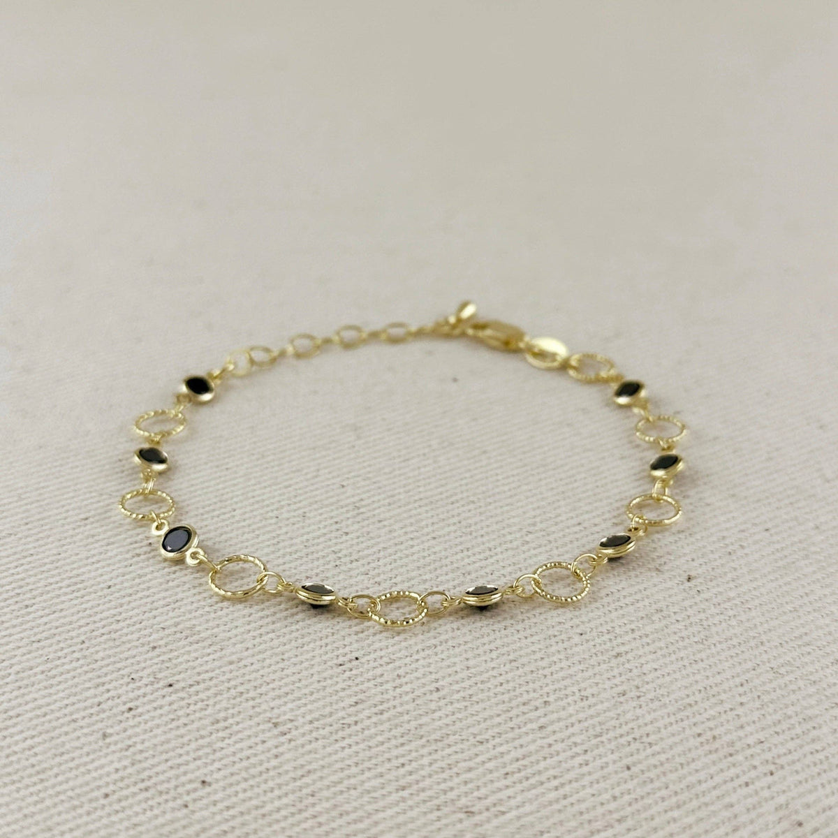 A delicate gold chain bracelet with small oval links and a few dark oval stone accents, displayed on a textured beige fabric background.