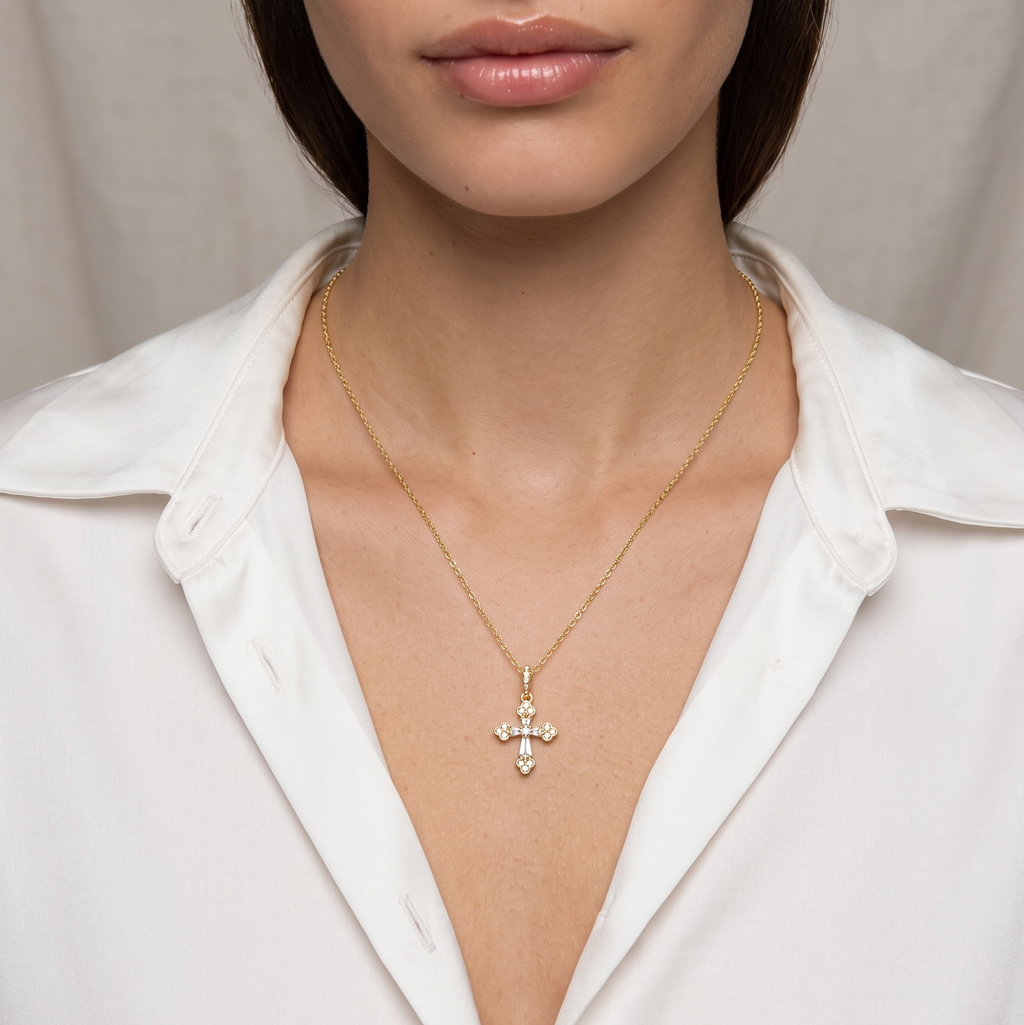 A close-up portrait of a model wearing a delicate gold cross pendant necklace with small clear stones, styled with a white collared shirt against a soft beige background.