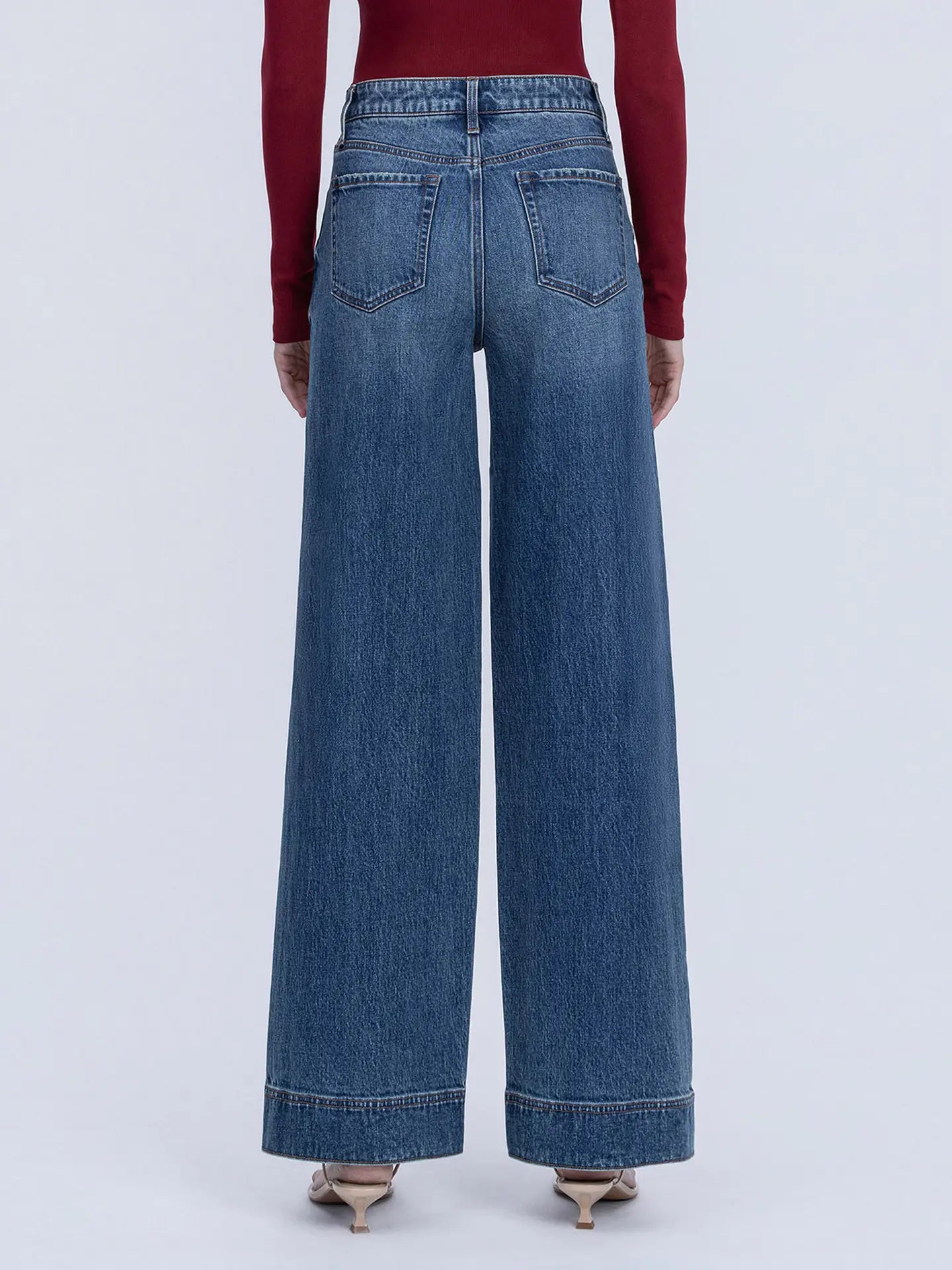 A person shown from the back wearing high-waisted, wide-leg blue denim jeans with a burgundy long-sleeve top and pointed-toe heels against a light gray studio background.