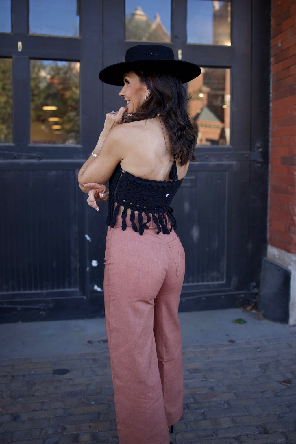 Woman wearing a black top with fringe and pink pants standing in front of a dark door.