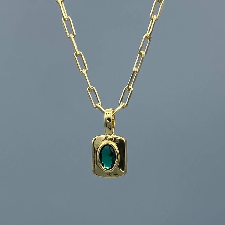 A gold paperclip chain necklace with a square bezel pendant featuring an oval-cut green gemstone, set against a solid slate blue background.