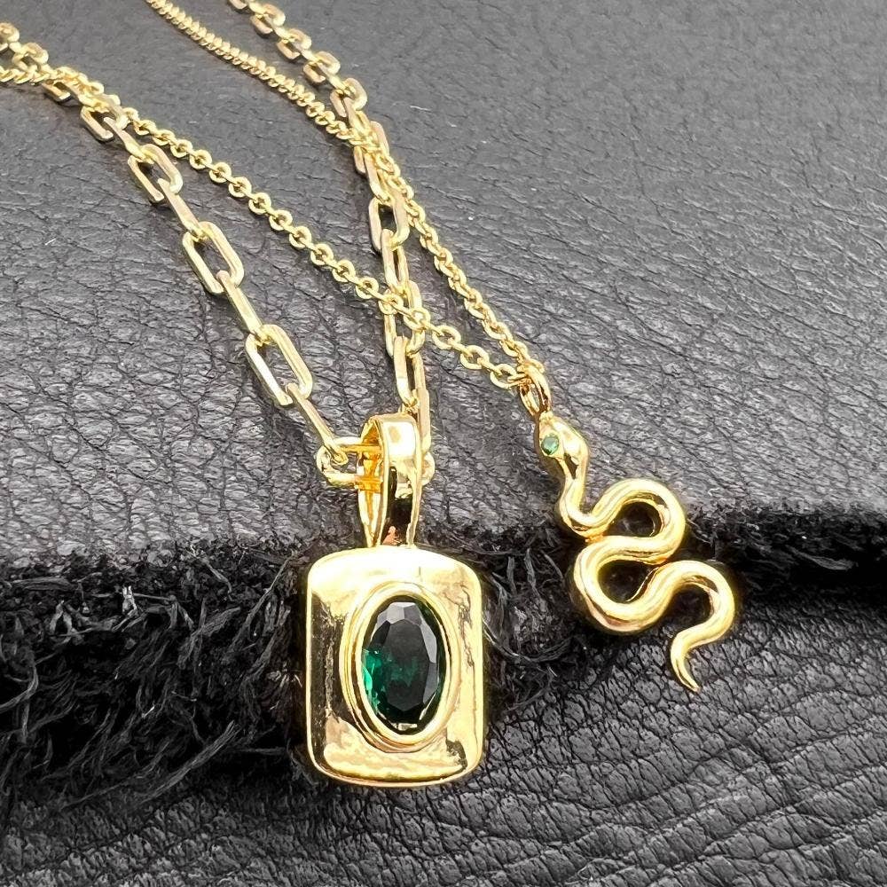 A gold necklace with two pendants displayed on a textured black surface: a square pendant with an oval green gemstone and a separate snake-shaped pendant, both on gold-tone chains.