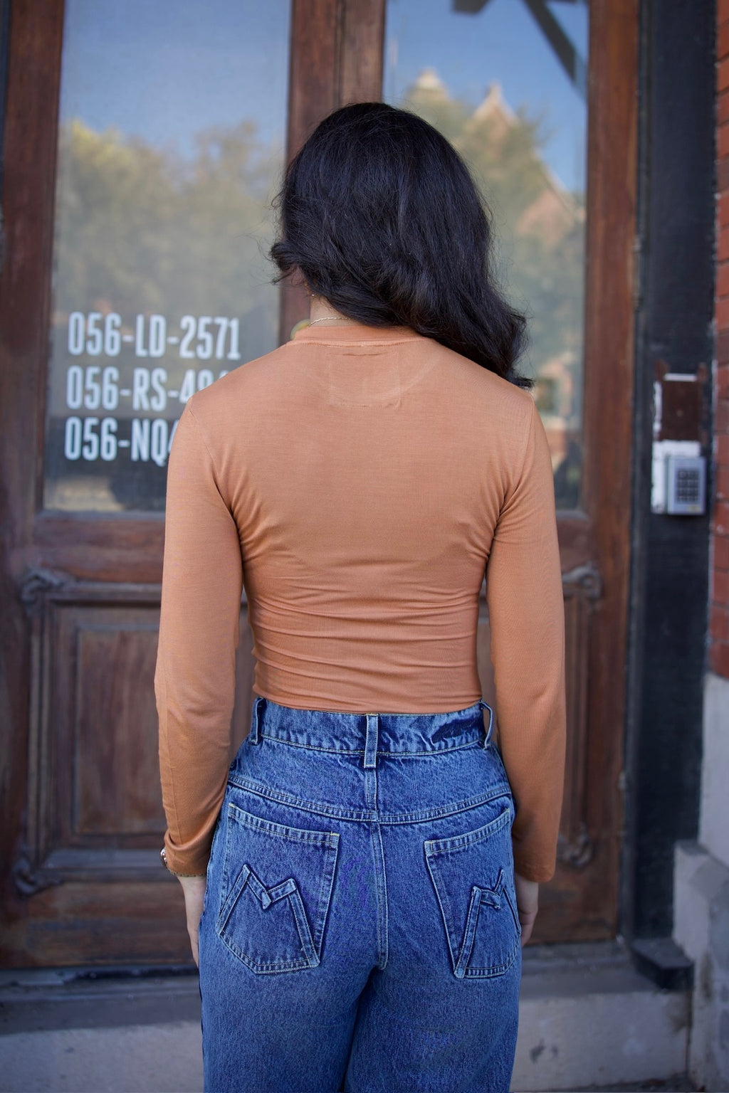 Person wearing a tan long-sleeve top and blue jeans standing in front of a glass door.