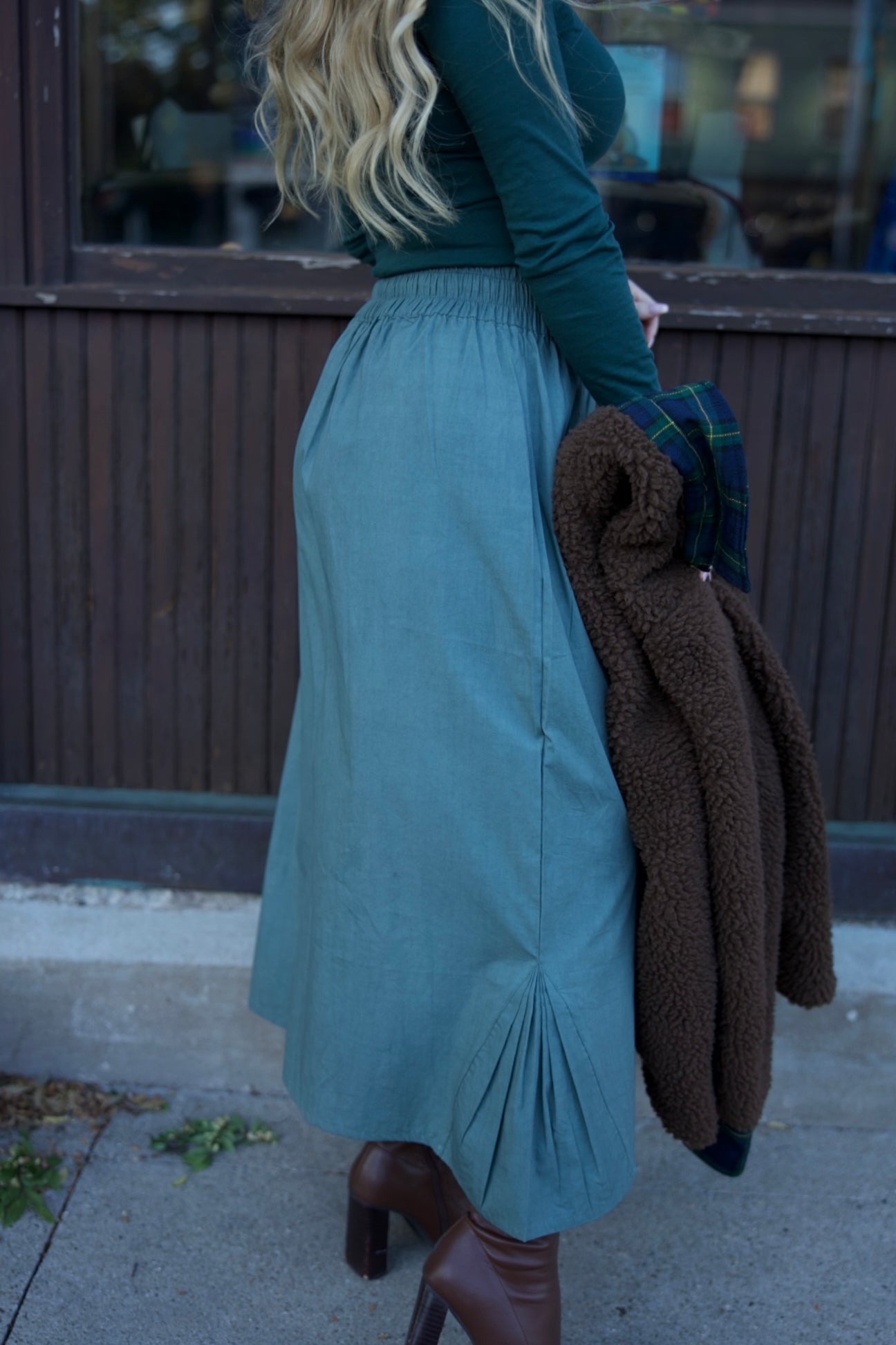 Person wearing a teal skirt and green top with a brown coat over shoulder, standing on a sidewalk.