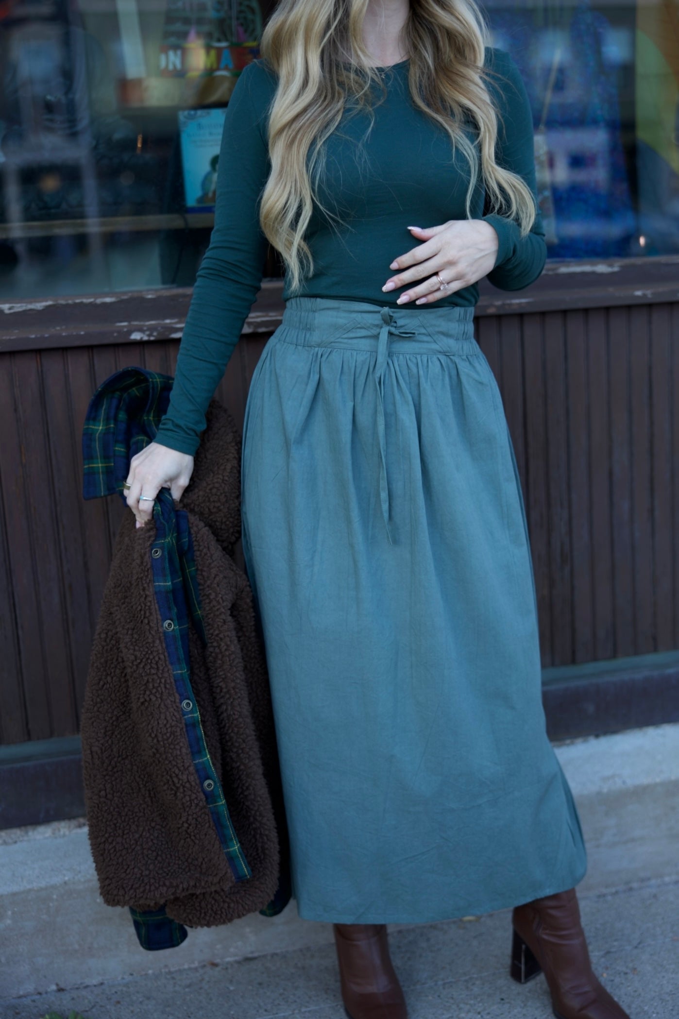 Woman in teal top and long skirt holding a brown coat against a blurred indoor background