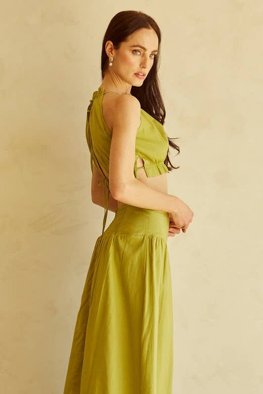 A woman modeling a sleeveless olive green two-piece set consisting of a halter-style crop top with side cutouts and a matching high-waisted pleated skirt, set against a textured beige background.