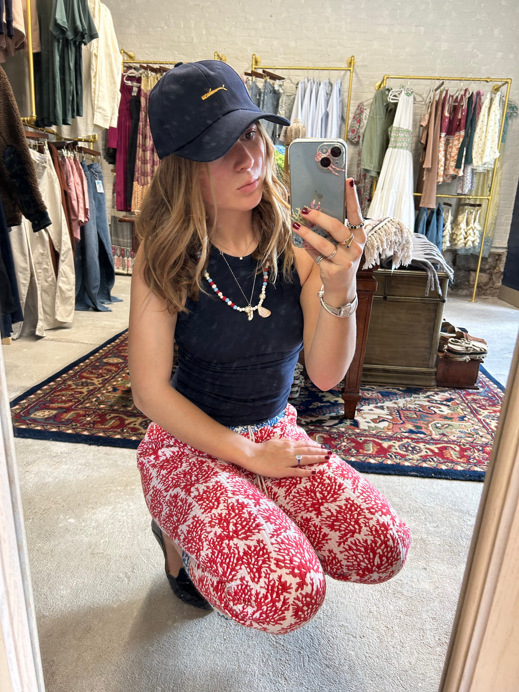 Woman taking a mirror selfie in a clothing store wearing a navy cap and red patterned pants.