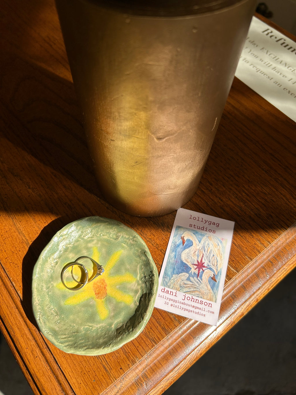 Small green dish with a yellow flower design on a wooden surface, next to a brass object and a card.