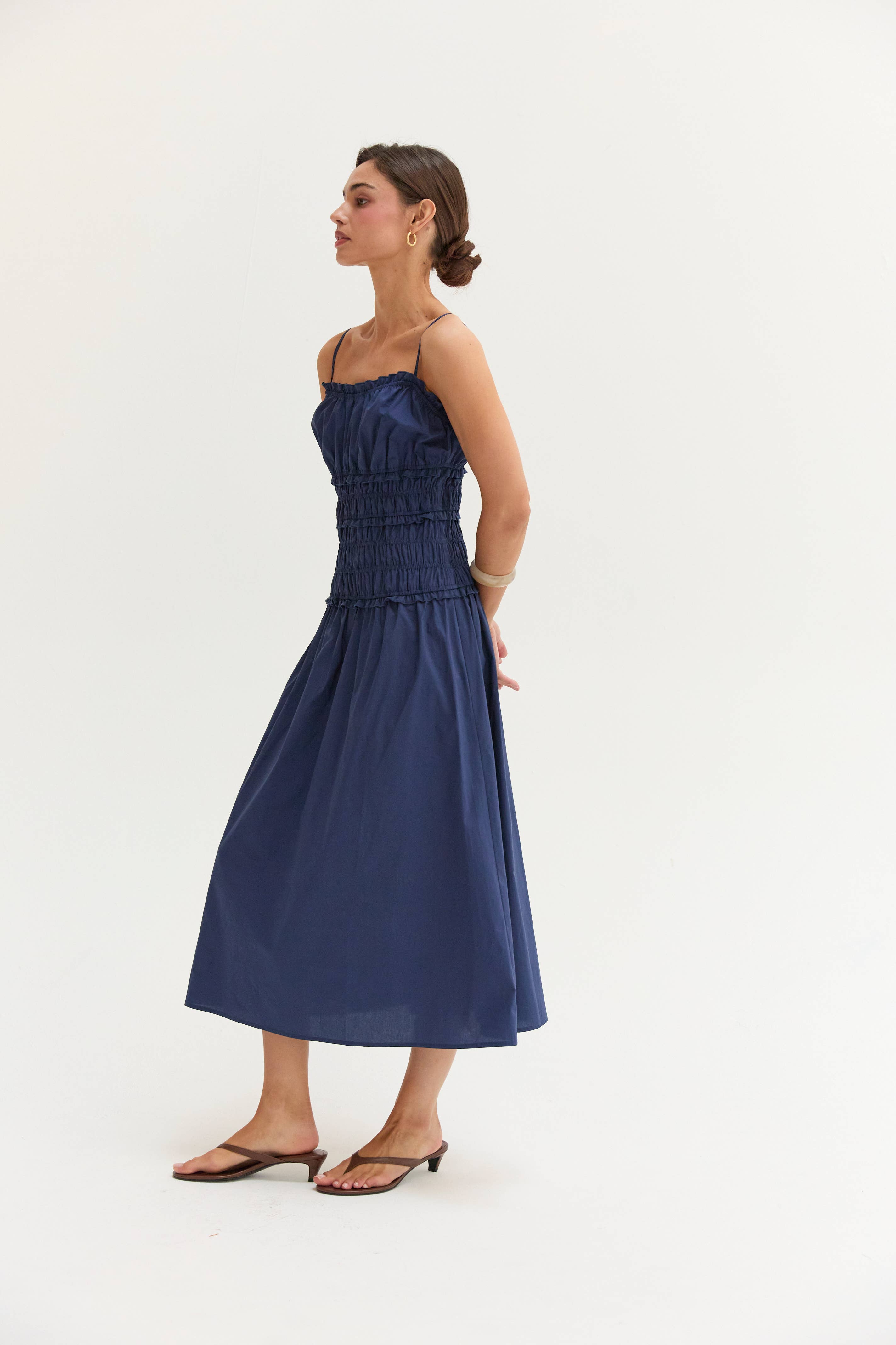 A woman modeling a navy blue midi dress with thin straps, a smocked bodice, and a flared skirt, paired with brown flat sandals against a plain white studio background.