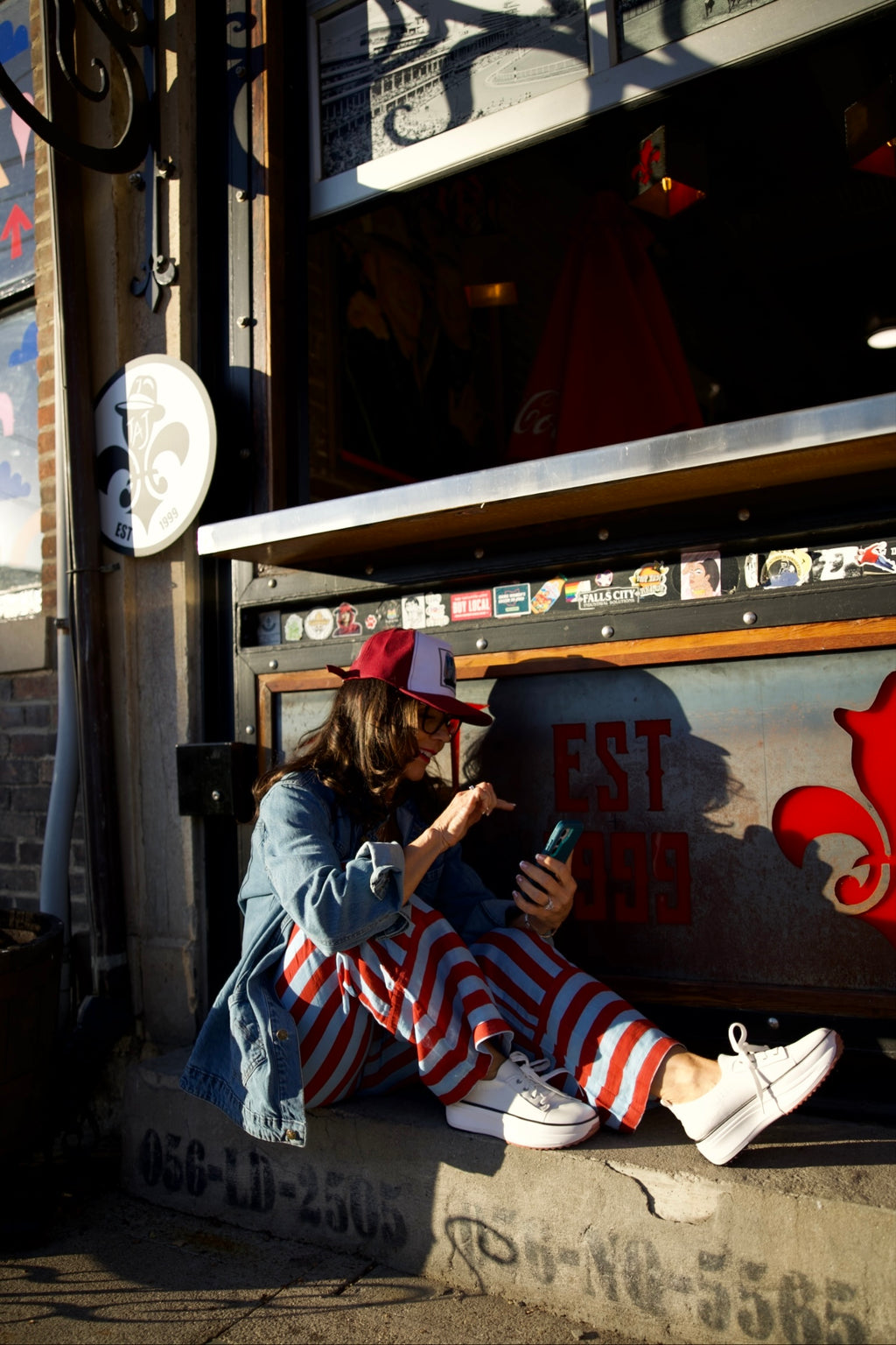 Person sitting on a bus stop bench using a phone, wearing a red cap and striped pants.