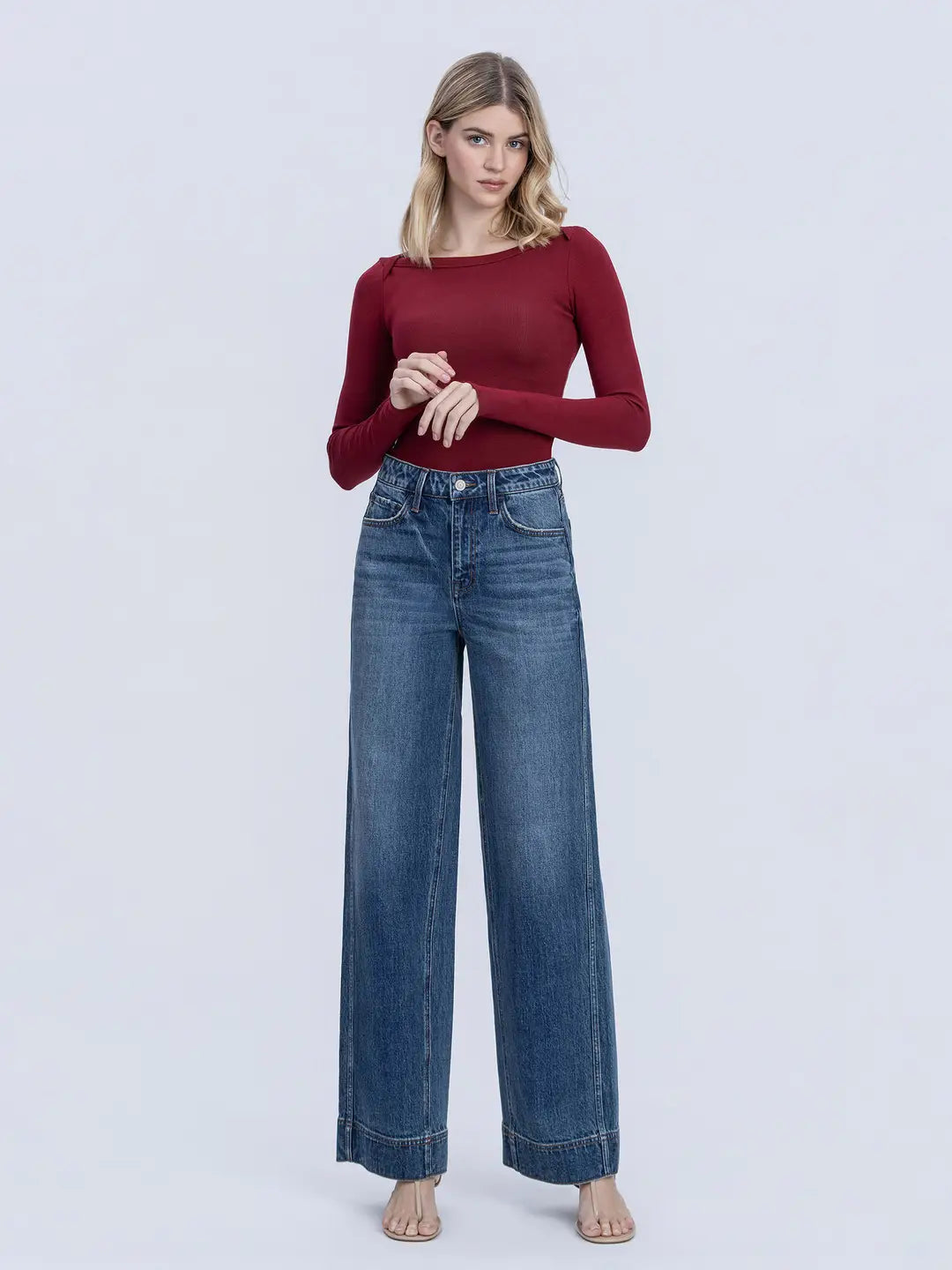 A woman modeling a long-sleeve burgundy knit top and high-waisted, wide-leg blue jeans against a light gray studio background.